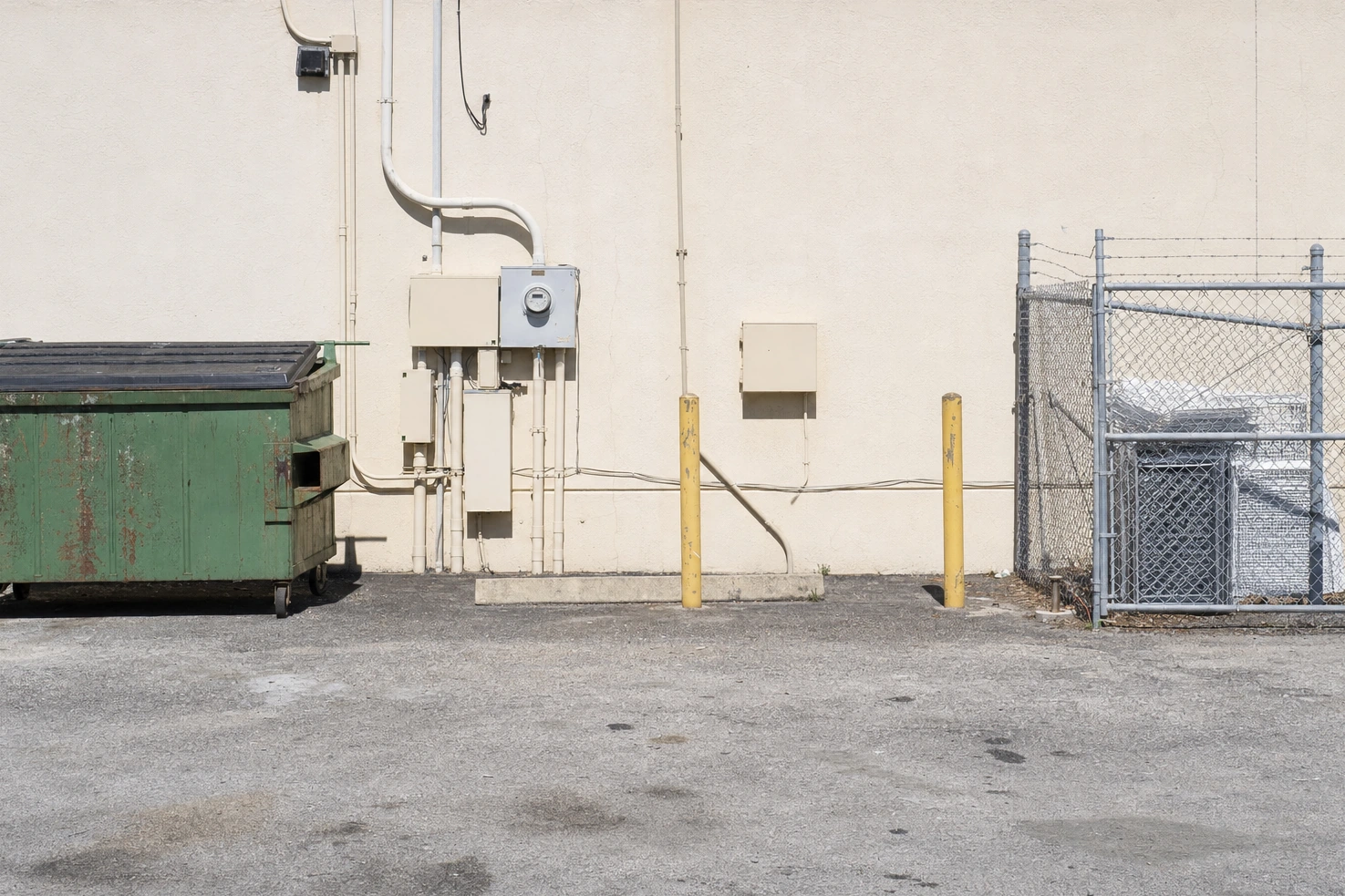 Our photograph of a service yard behind a commercial building at midday: dumpsters, exposed conduit, a chain-link fence and a blank stucco wall.