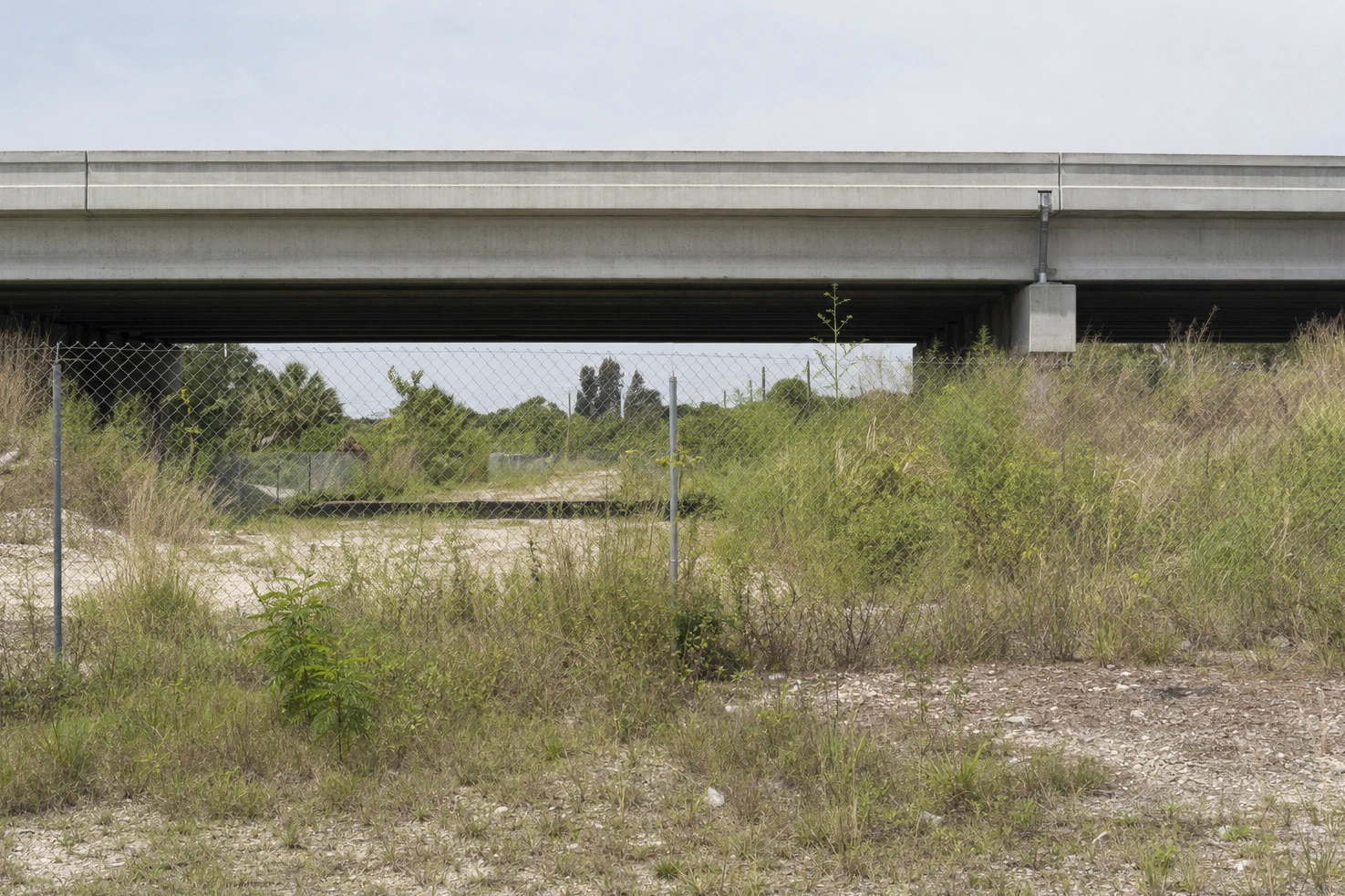 Our photograph of a concrete overpass crossing the frame horizontally in flat midday light, with scrub and fencing underneath it.
