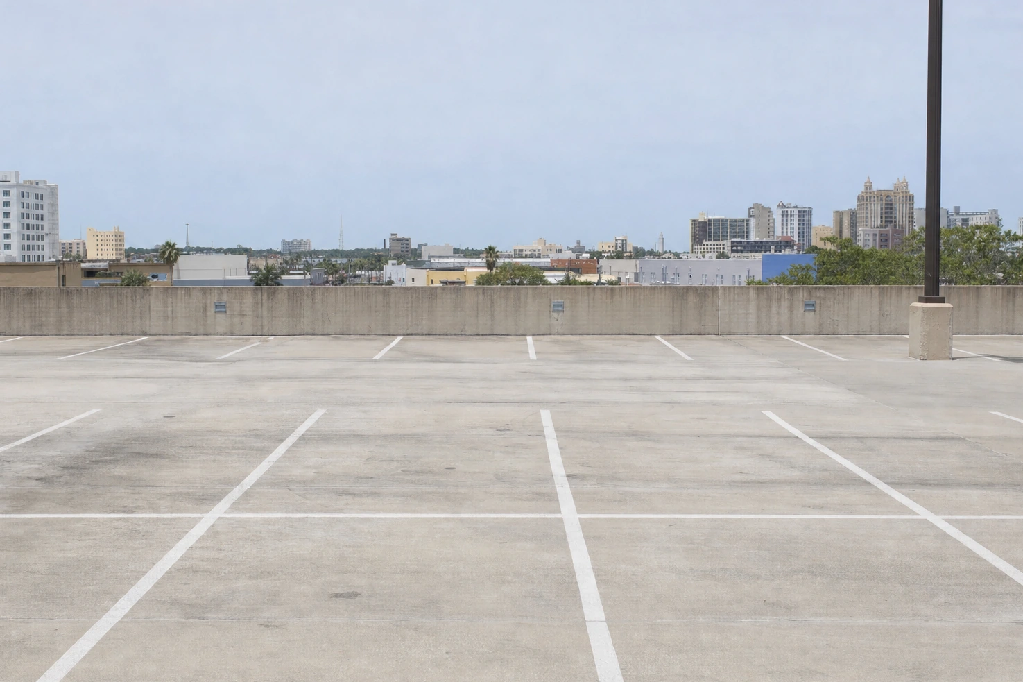 Our picture of the open top deck of a multi-storey car park at midday: painted bays, a low perimeter wall, the city beyond it under flat light.