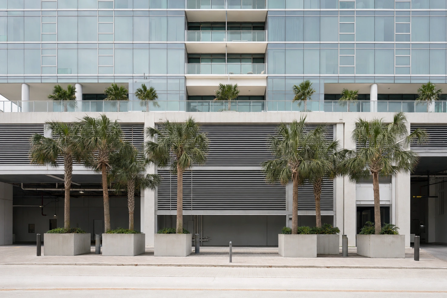 A picture we took of the base of a glass residential tower where it meets its parking podium, photographed frontally at midday, palms in concrete planters.