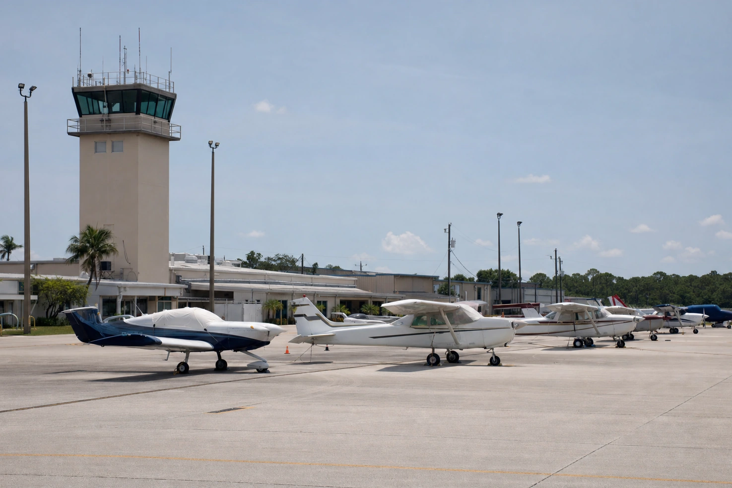 Our picture of a regional airfield apron at midday: light aircraft parked in a row, a control tower behind them, heat coming off the concrete.