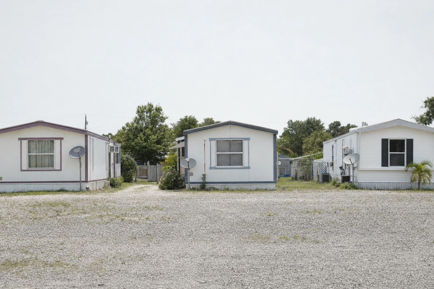 A row of single-wide trailers we photographed frontally at midday: gravel underfoot, satellite dishes on the ends, flat light.