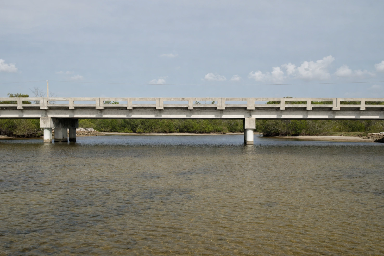 Our photograph of a low concrete bridge span crossing the frame horizontally over shallow water in flat midday light.