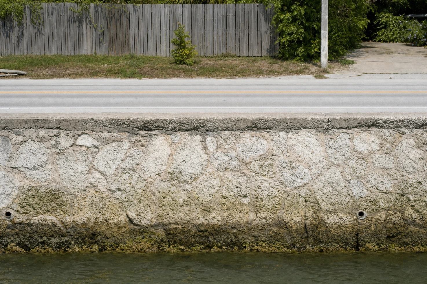A limestone seawall we photographed running the width of the frame at midday, water on one side of it and road on the other.