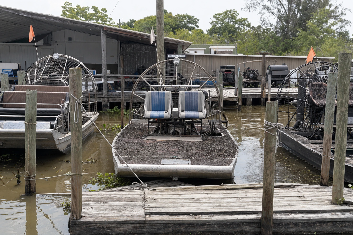 Airboats tied up at a working dock, photographed by us frontally at midday over mud and boards.
