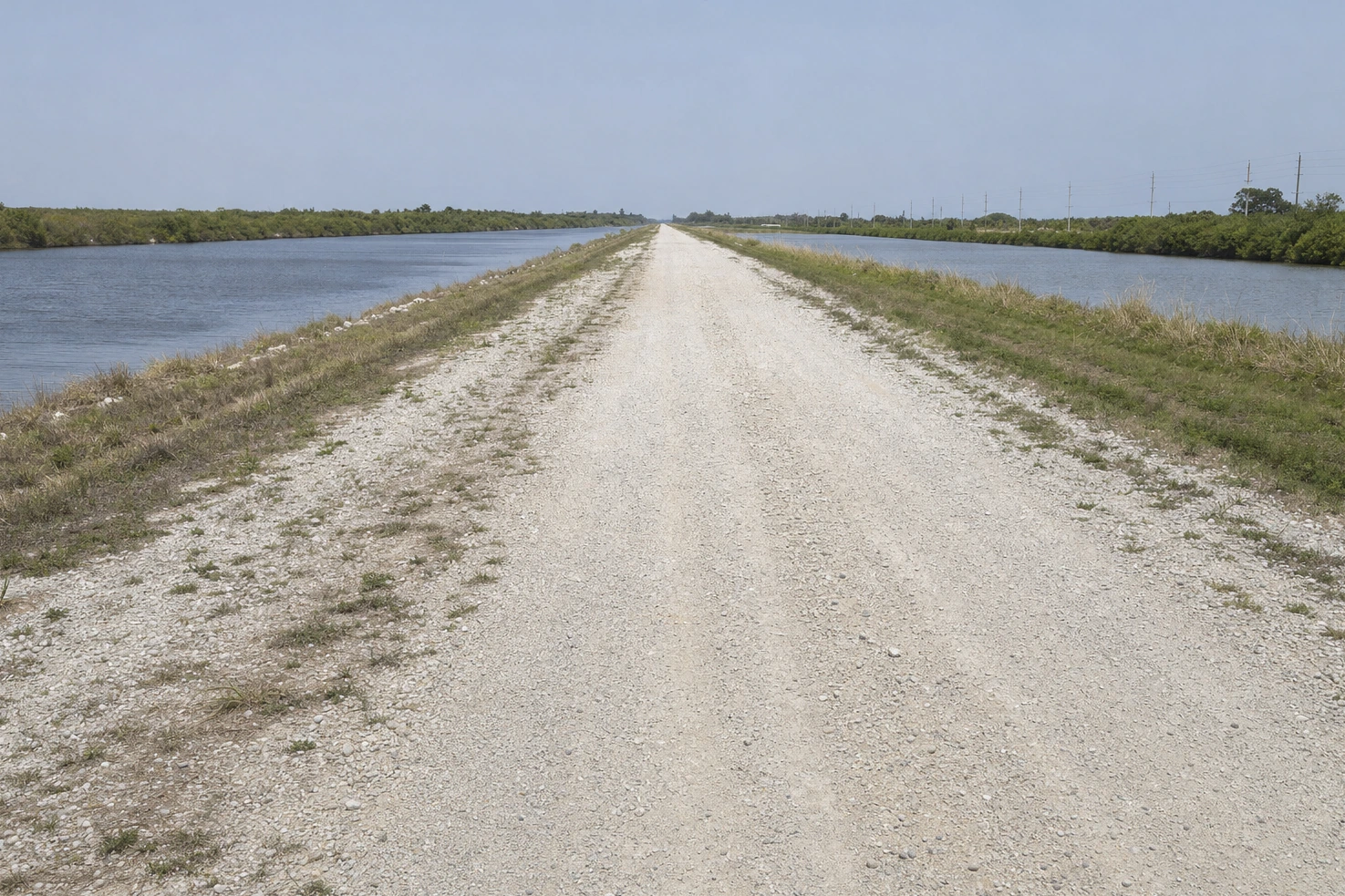 Our picture of a straight gravel levee road running to the horizon between two water channels at midday.